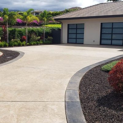 A beautifully designed home with a wide, smooth concrete driveway bordered by dark trim and lush tropical landscaping, a perfect example of expert Concrete Driveways Services.