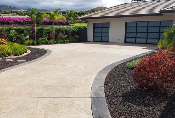 A beautifully designed home with a wide, smooth concrete driveway bordered by dark trim and lush tropical landscaping, a perfect example of expert Concrete Driveways Services.