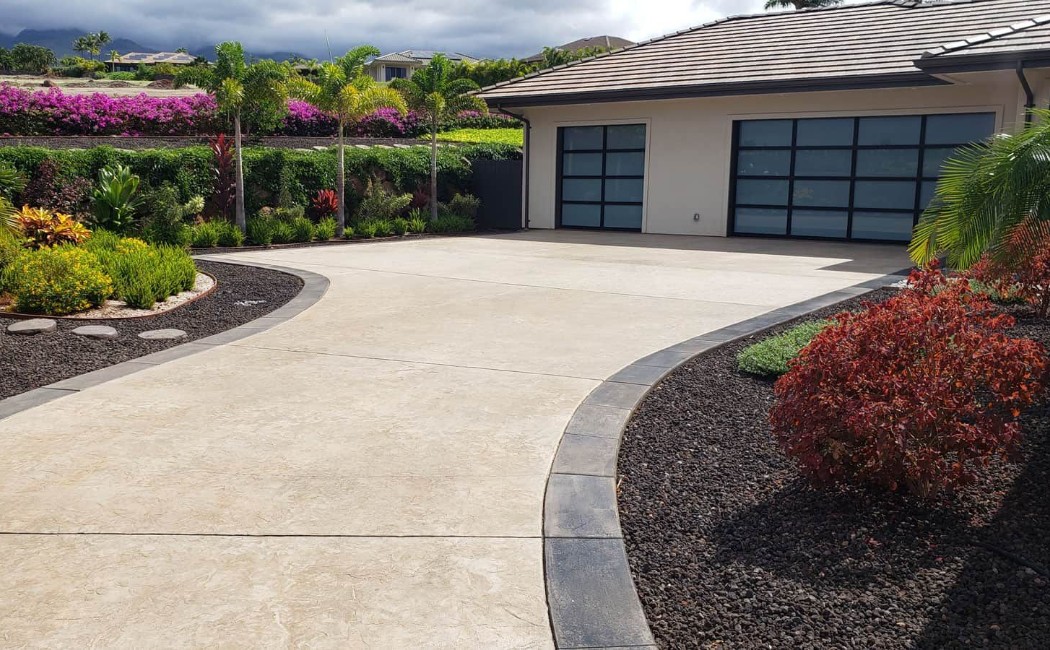 A beautifully designed home with a wide, smooth concrete driveway bordered by dark trim and lush tropical landscaping, a perfect example of expert Concrete Driveways Services.