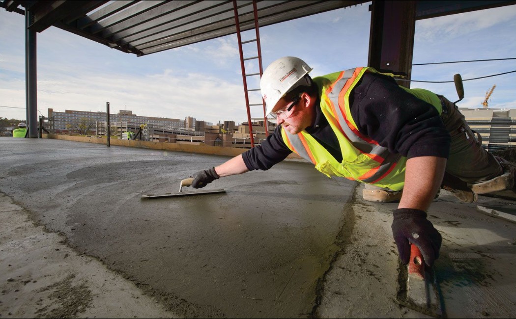 A construction worker from a Concrete Work Company expertly smooths freshly poured concrete on a high-rise building floor, showcasing precision and craftsmanship on the job site.