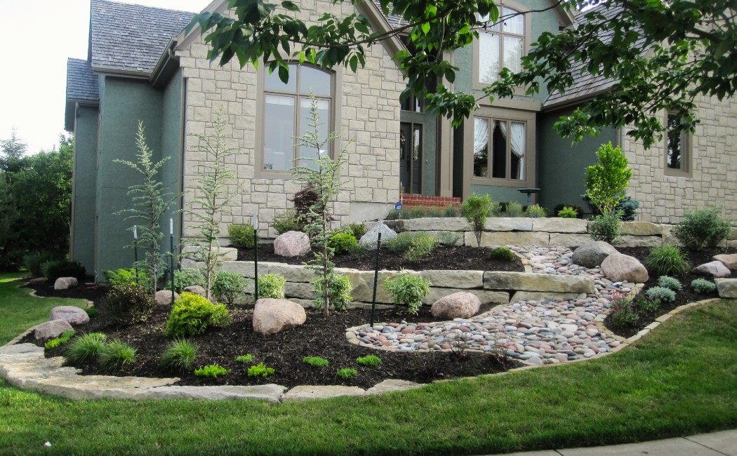 The image features a well-designed landscape with tiered stone flower beds, decorative rocks, and carefully placed plants, demonstrating professional landscaping techniques that enhance the home's curb appeal.