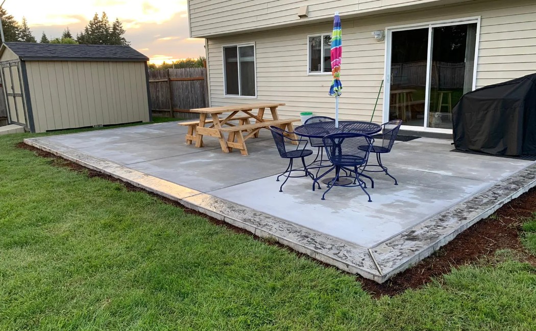 A spacious backyard concrete patio with outdoor seating, picnic table, and umbrella beside a residential home, showcasing professional Concrete Patios Services for durable and modern outdoor living spaces.
